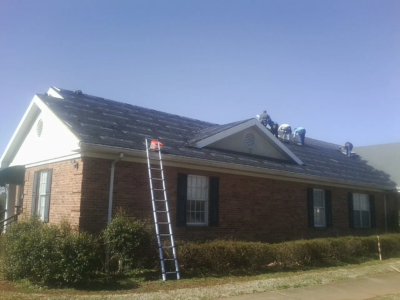 Crew tearing shingles off a brick ranch roof with a ladder set — Metal Roof Repair work by our crew in Fairhaven