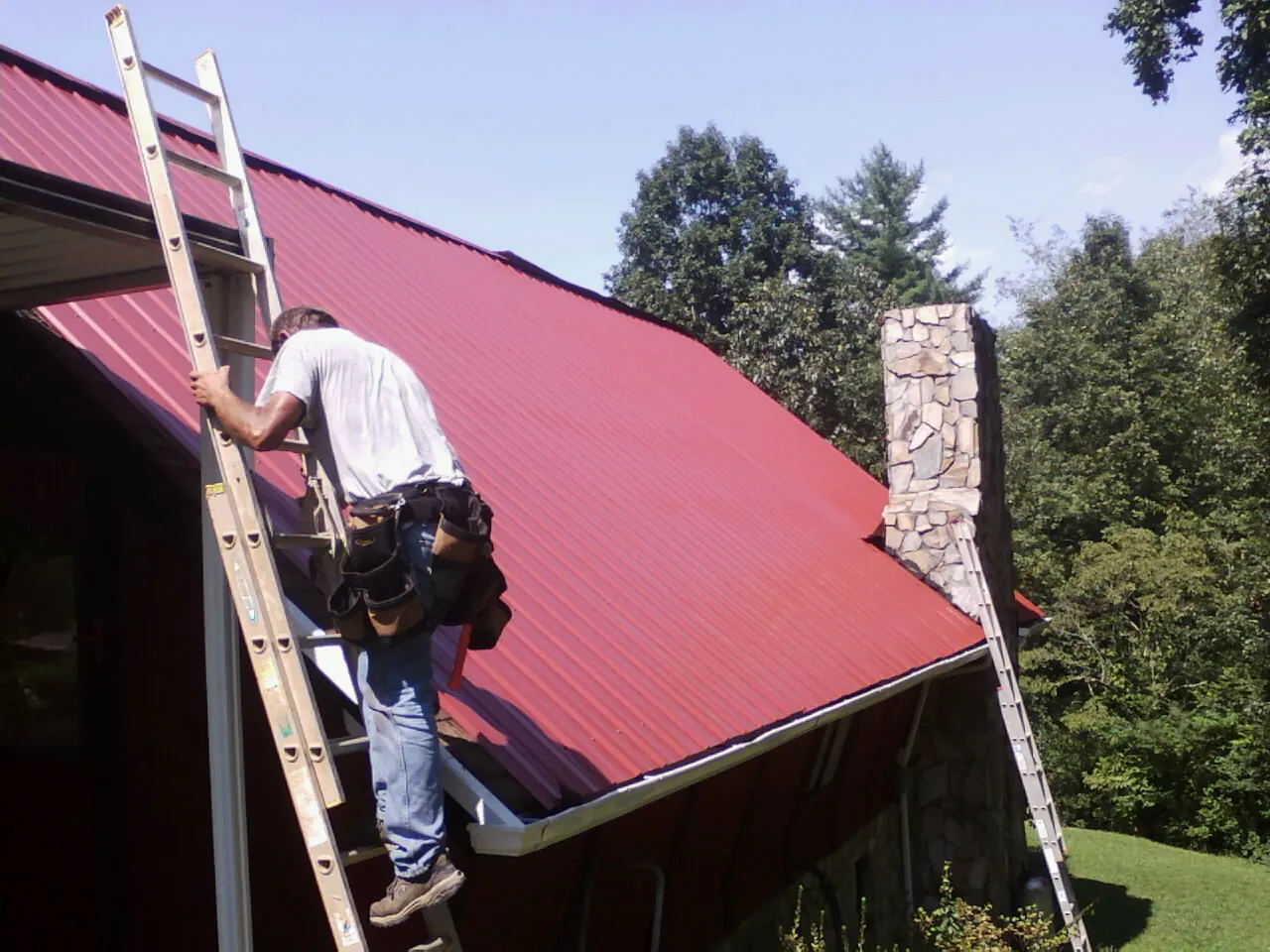 Roofer on a ladder beside a red metal roof and stone chimney — Soffit & Fascia Repair by Roofing Brothers in Fairhaven
